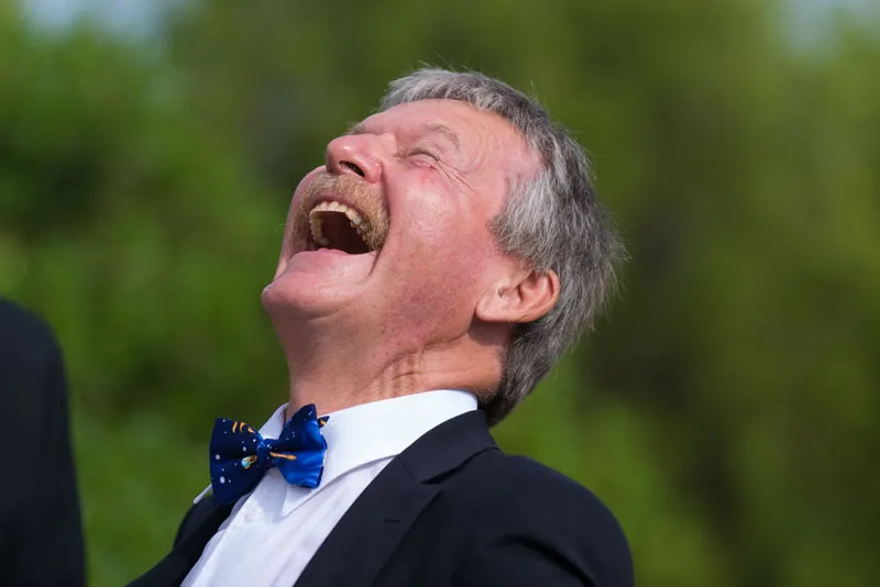 Older man in a dark suit and blue bow tie laughing joyfully with his head tilted back against a green blurred background.