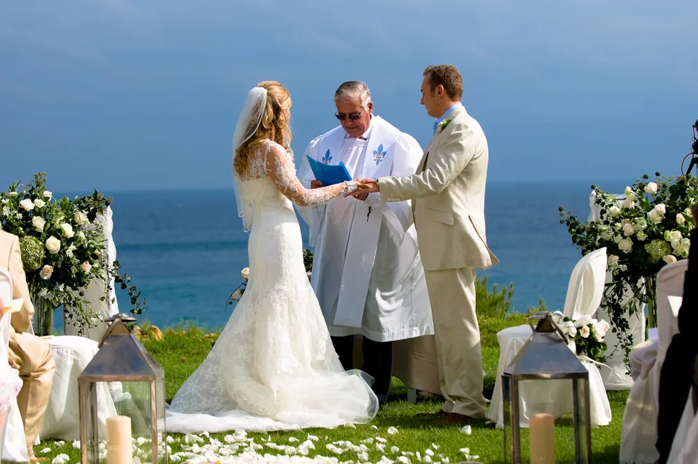Bride and groom holding hands at outdoor wedding ceremony by the ocean, with officiant in white robe.