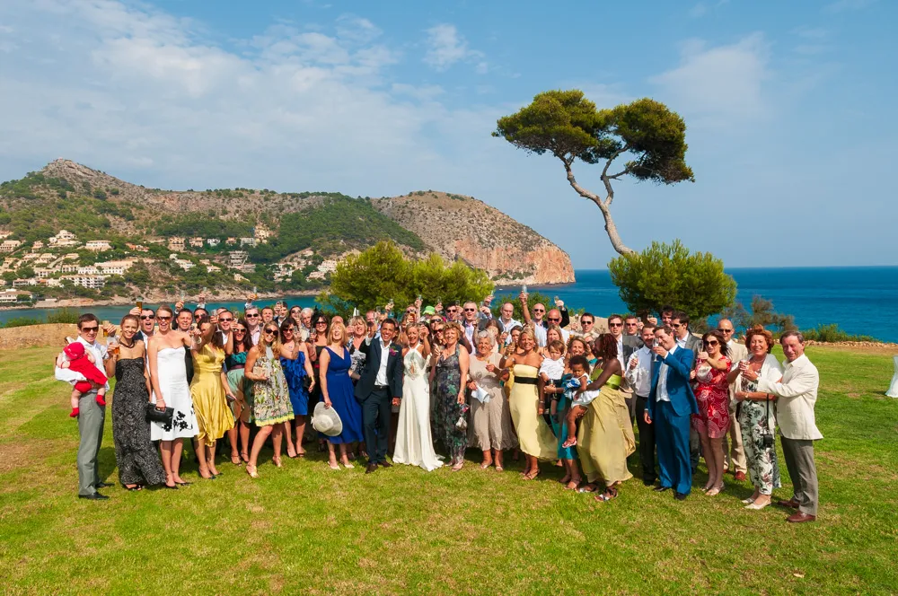 Large group of people dressed in formal attire celebrating outdoors on grass, with a scenic coastal hillside and blue sea in the background.