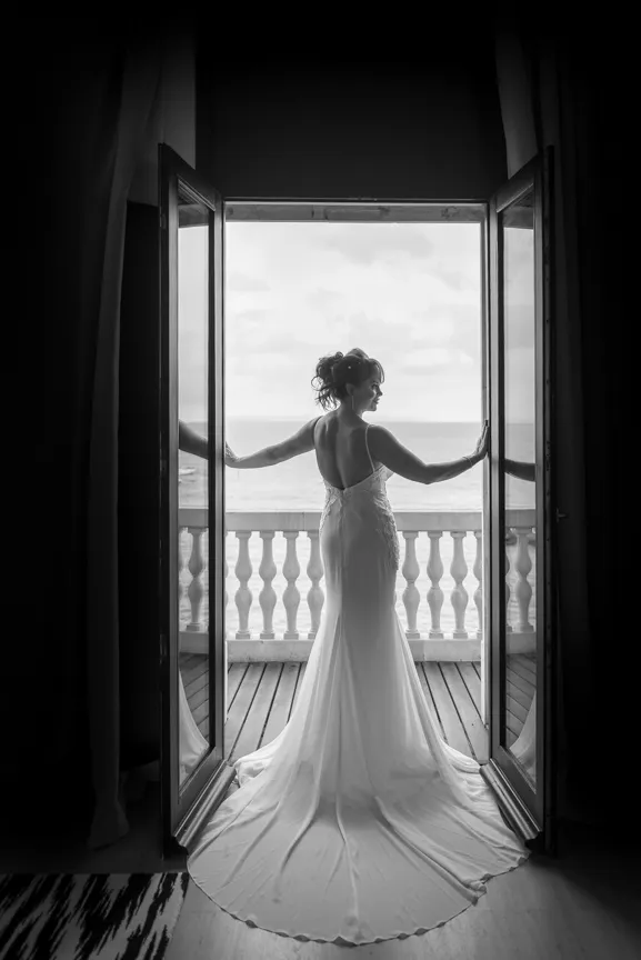 Bride in a long wedding dress standing in an open doorway facing a balcony with ocean view.