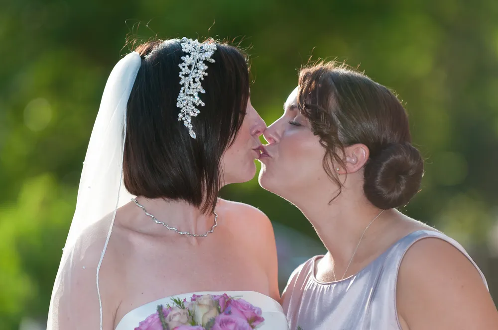 Two women with elegant hairstyles and dresses sharing a kiss outdoors, one wearing a bridal veil and holding a bouquet.