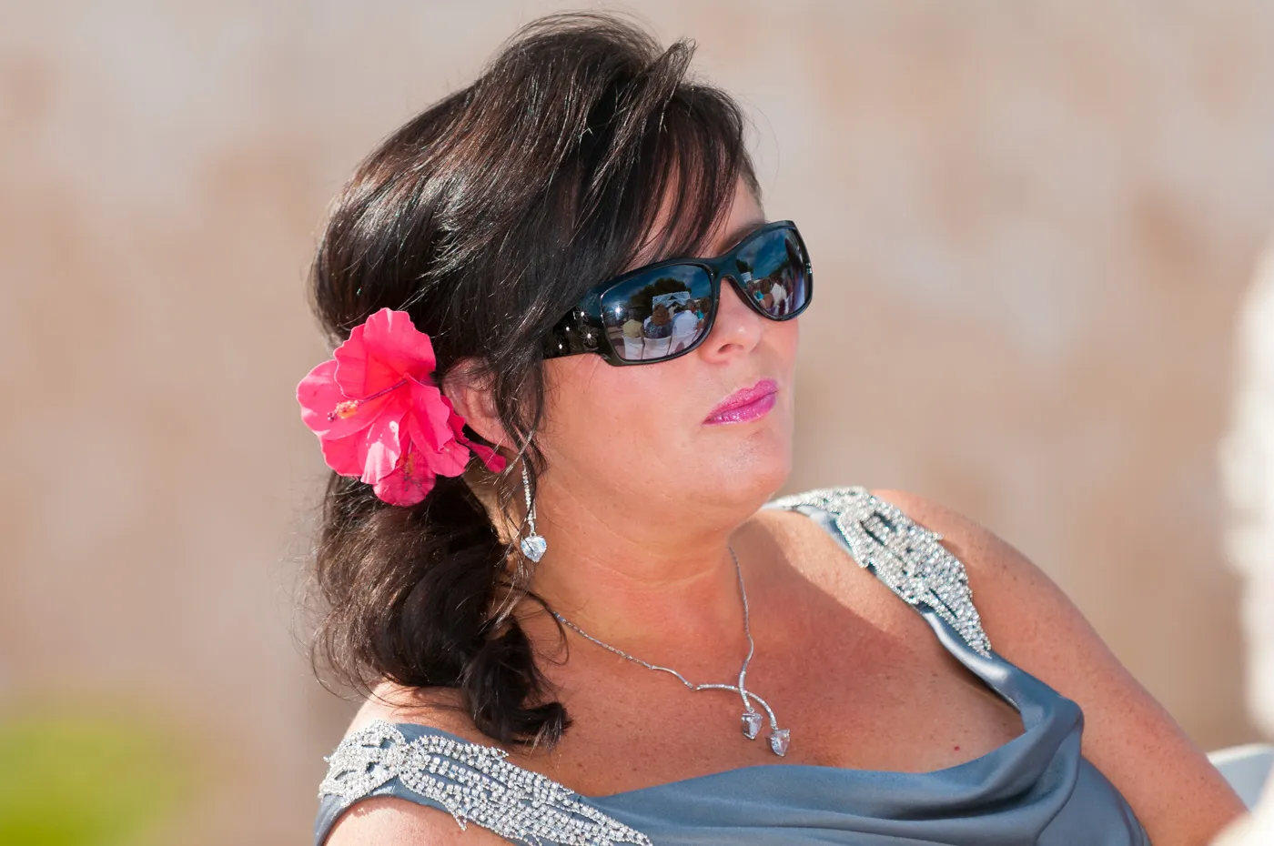 Woman with dark hair wearing black sunglasses, a silver dress with rhinestone detailing, a pink flower in her hair, and a silver necklace and earrings.