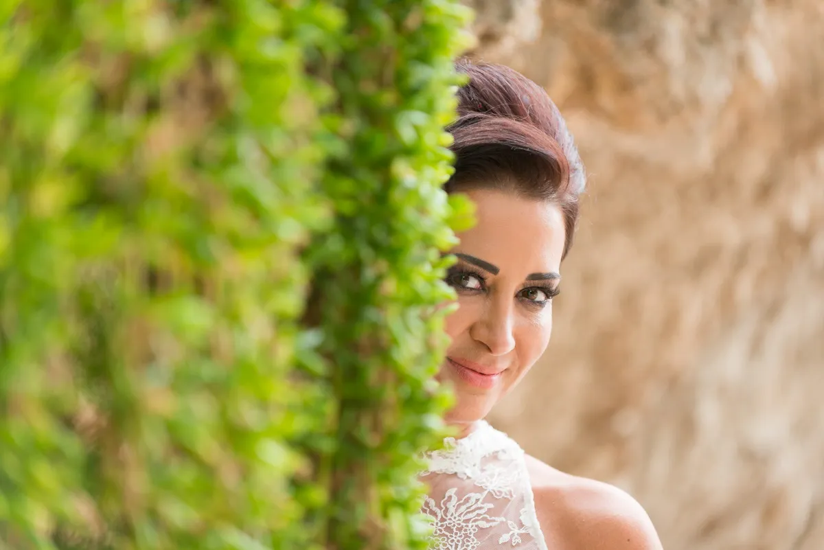 Woman in white lace dress smiling while partially hidden behind green foliage with a blurred brown background.