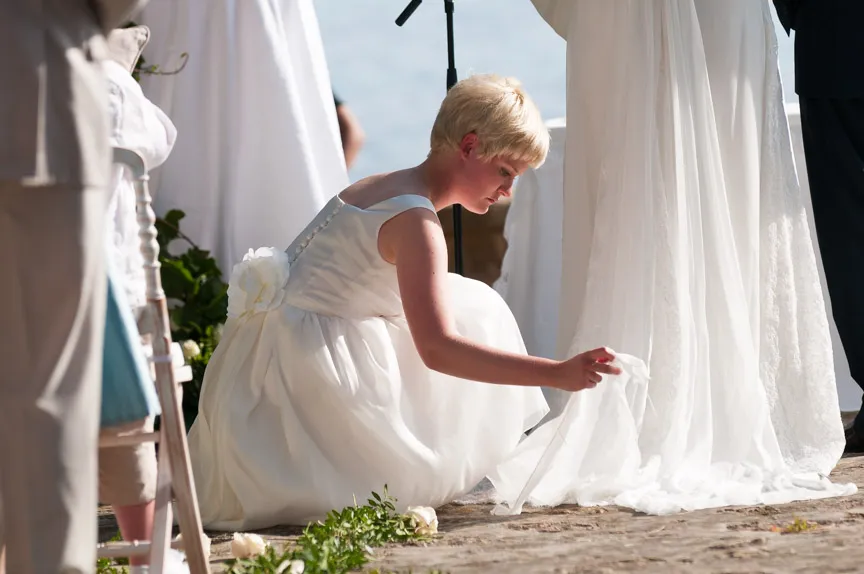 Young flower girl in a white dress kneeling and adjusting the train of a bride's wedding gown outdoors.