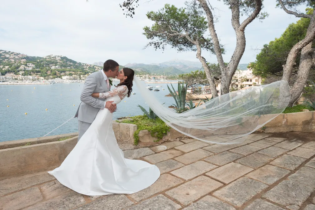 Bride and groom kissing near a waterfront with mountains and boats in the background, bride's veil flowing in the wind.