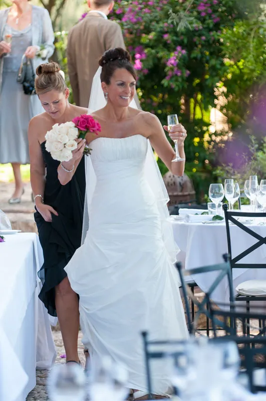 Bride in white strapless gown holding a glass of champagne and bouquet, followed by bridesmaid in black dress holding white bouquet, walking between dining tables outdoors.
