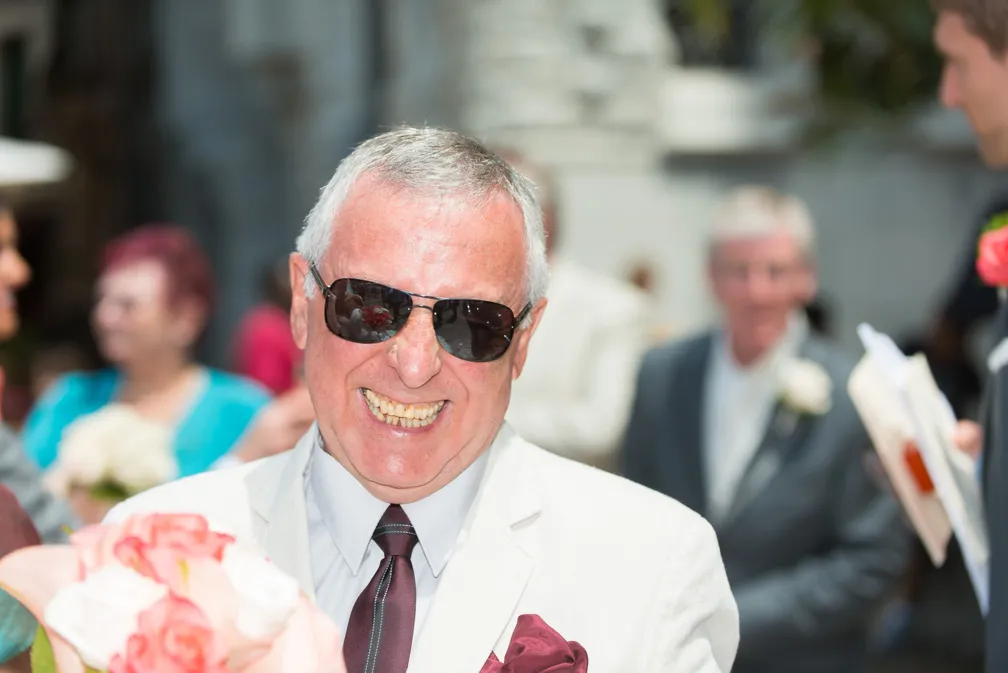 Smiling elderly man wearing sunglasses, a white suit, and a burgundy tie holds a bouquet of pink and white flowers at an outdoor event.