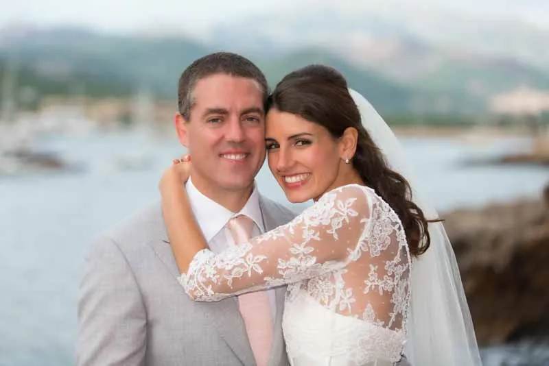 Smiling bride in a lace wedding gown embraces groom in light gray suit with a body of water and mountains in the background.