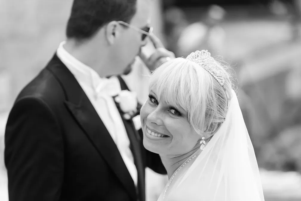 Smiling bride wearing a tiara and veil looks at the camera while groom in a tuxedo adjusts his sunglasses in the background.