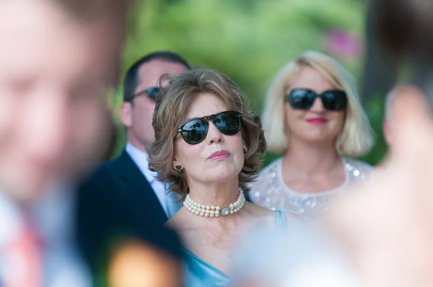 Two women wearing sunglasses at an outdoor event, one with a pearl necklace and blue dress, the other with blonde hair and a sparkly top.