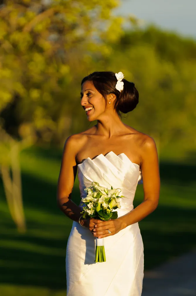Smiling bride in a strapless white wedding dress holding a bouquet of white flowers outdoors.