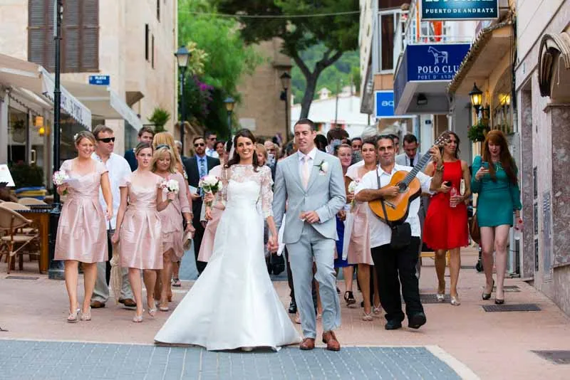 Bride in a white gown and groom in a light gray suit walking hand in hand with bridesmaids in pale pink dresses and a man playing guitar on a street.