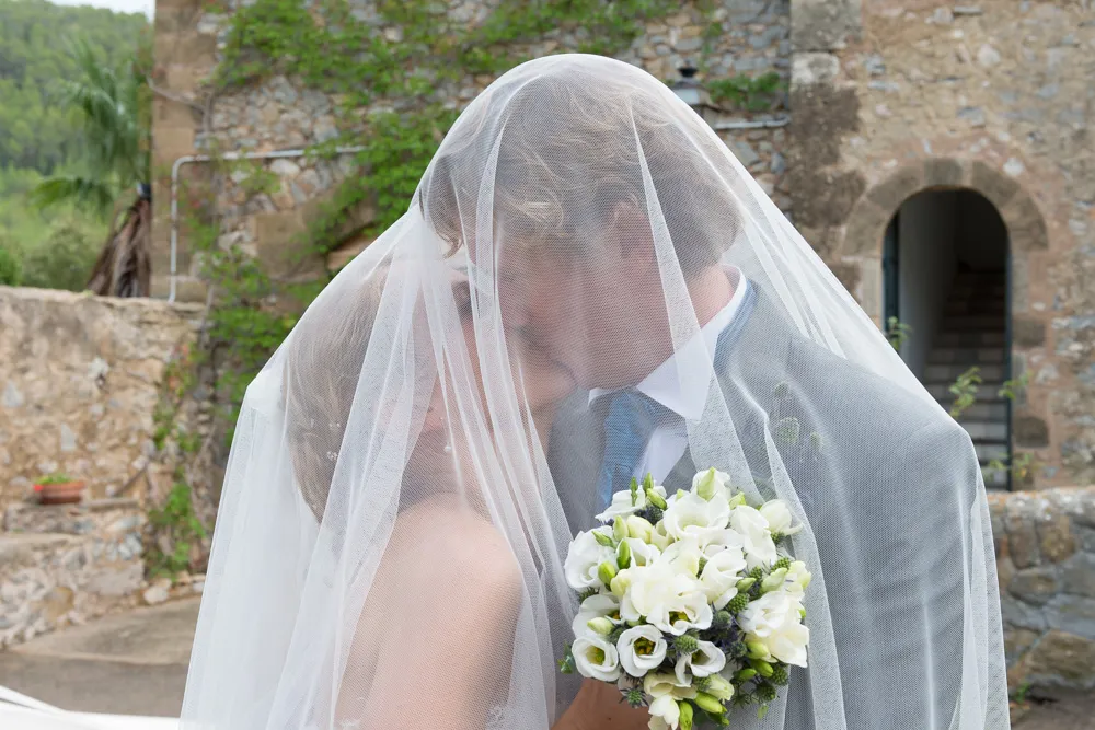 Bride and groom embracing under a wedding veil with a stone building background, groom holding a bouquet of white flowers.