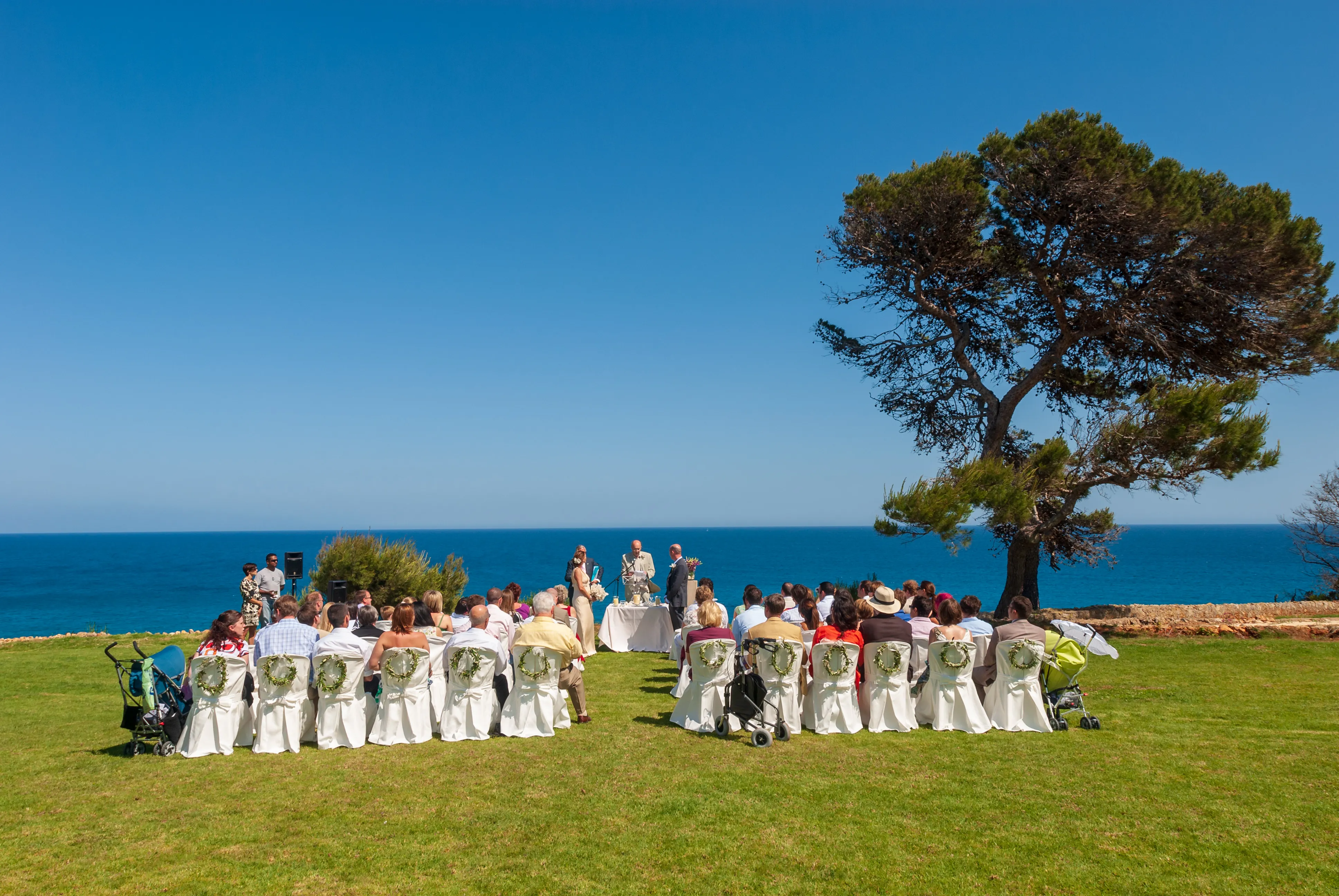 Outdoor wedding ceremony by the sea with guests seated on white chairs decorated with greenery.