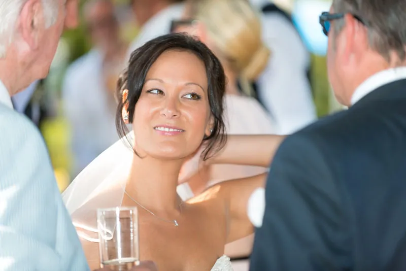 Smiling bride with dark hair and veil chatting with two men at a social event.