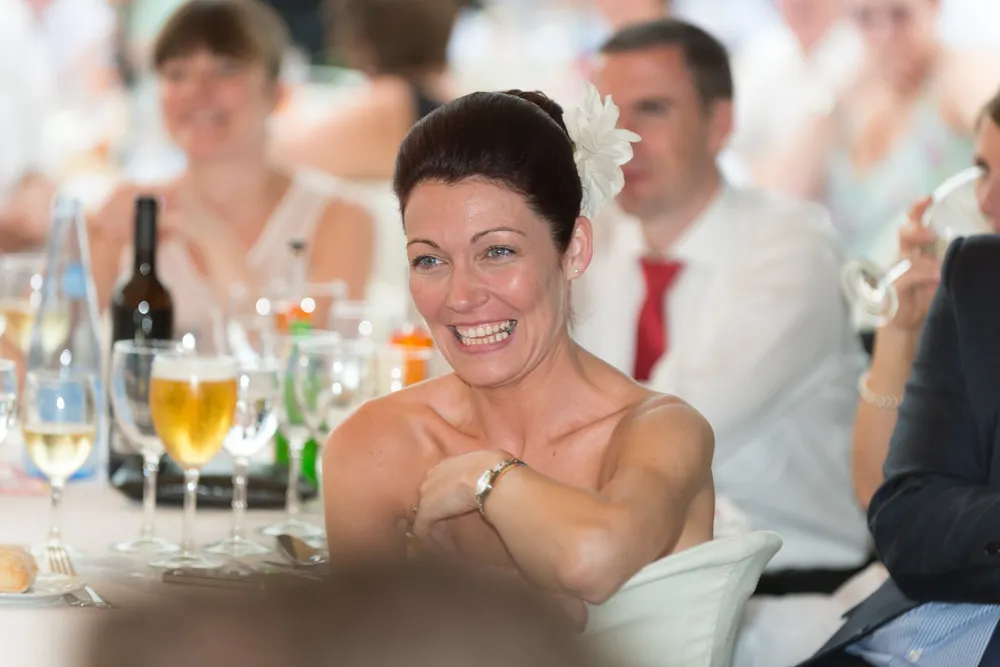 Smiling woman with a white flower in her hair sitting at a table set with glasses and bottles, surrounded by guests.