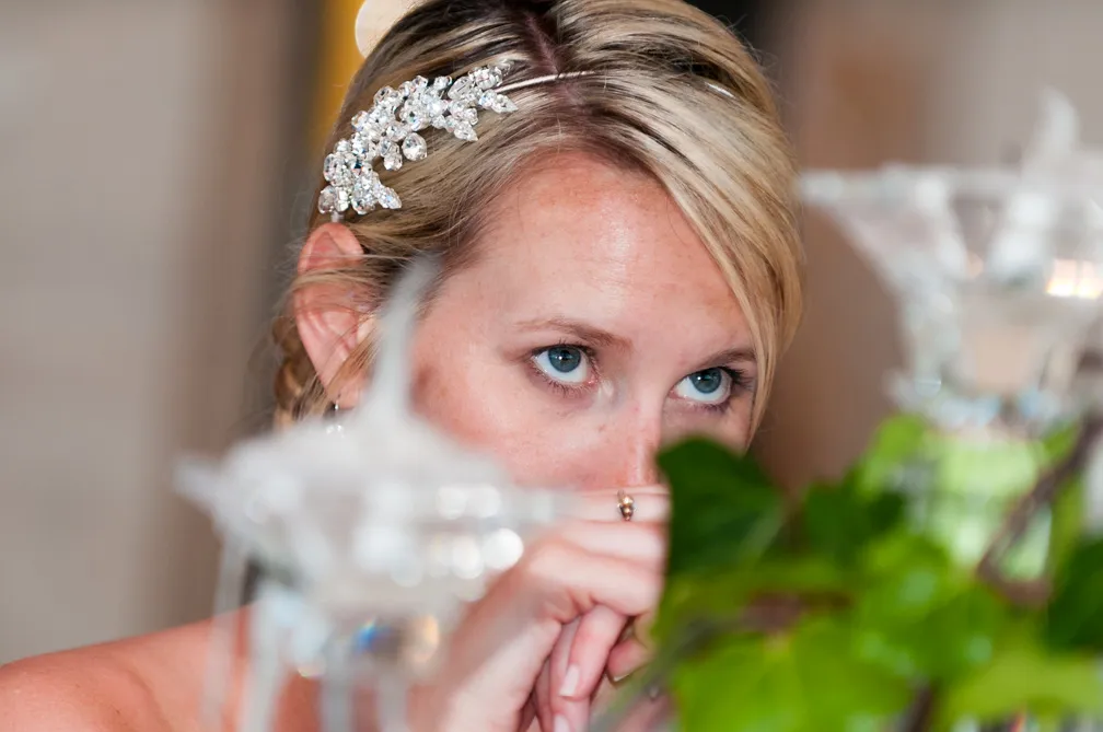 Close-up of a woman with blue eyes and a jeweled hair accessory, partially obscured by glass decorations and greenery.
