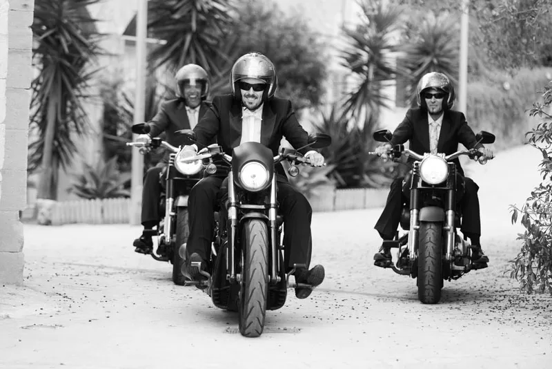 Three men in tuxedos and helmets riding motorcycles down a street with plants on both sides.