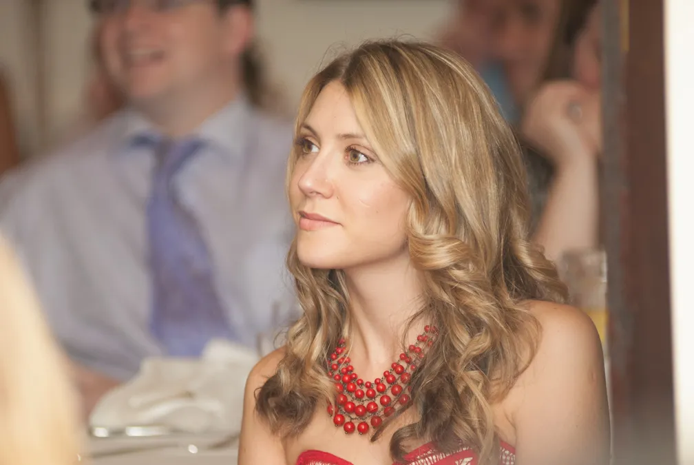 Blonde woman with curly hair wearing a red strapless dress and red beaded necklace, looking to the side.