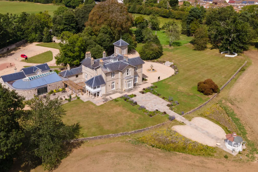 A large historic stone mansion with multiple peaked roofs, surrounded by green lawns, trees, a driveway with a red car, and a small white gazebo.