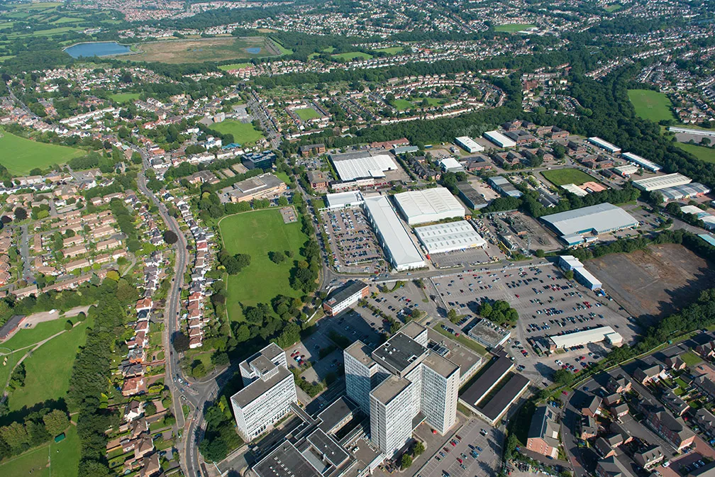 Aerial view of a suburban area with residential houses, green fields, several large commercial buildings, and parking lots.