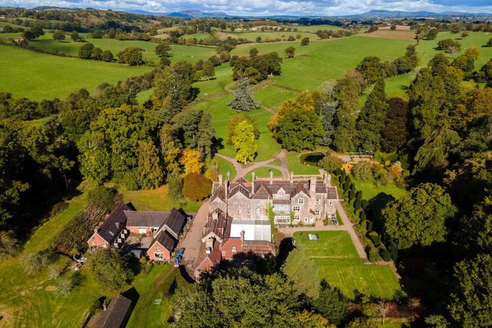 Aerial view of a large historic mansion surrounded by gardens, trees, and expansive green fields under a partly cloudy sky.