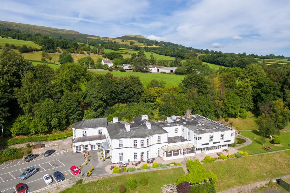 A large white building with a parking area, surrounded by green trees and rolling hills under a partly cloudy sky.