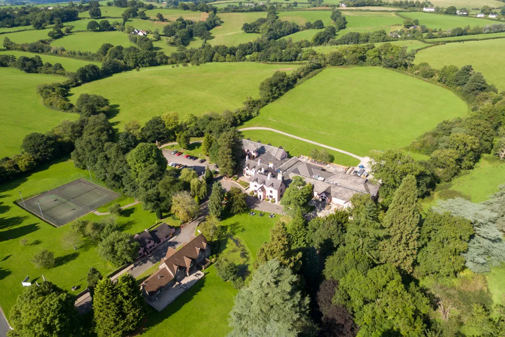 Aerial view of a countryside estate with large historic buildings surrounded by green fields, trees, and a tennis court.