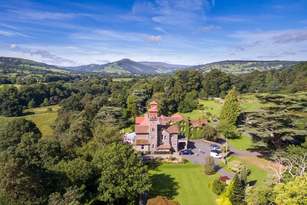 A stone building with red-tiled roofs surrounded by green trees and lawn, set in a scenic valley with rolling hills in the background under a blue sky.