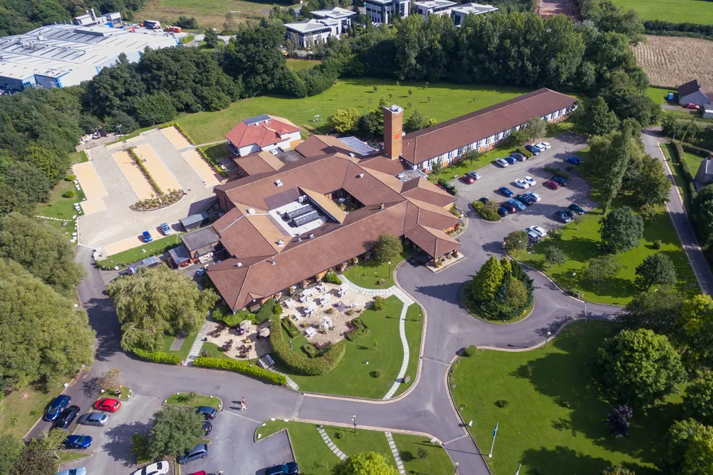 Aerial view of a large building complex with brown roofs surrounded by green lawns, parking lots, and trees.