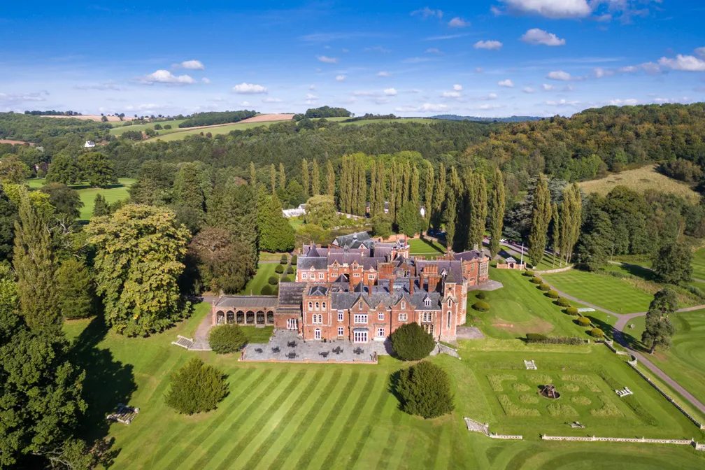 Aerial view of a large historic red-brick mansion surrounded by manicured lawns, trees, and landscaped gardens under a blue sky.