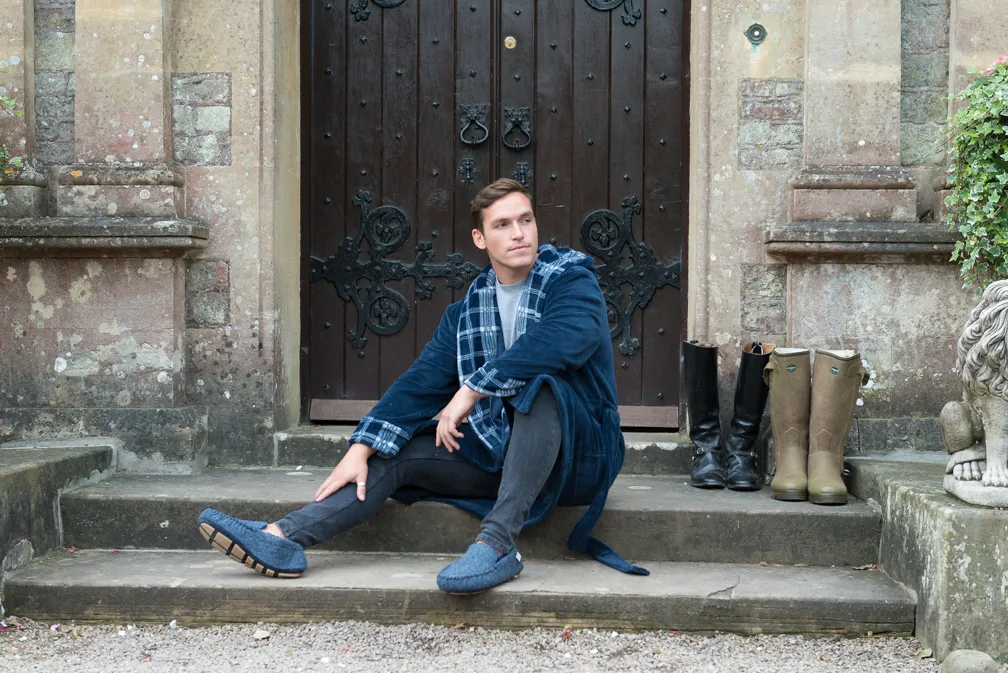 Man in blue robe and slippers sitting on stone steps in front of a large wooden door with pairs of boots beside him.