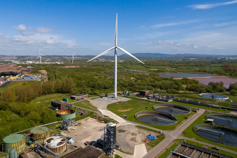 Aerial view of a wastewater treatment facility with multiple circular settling tanks and three wind turbines surrounded by green fields under a partly cloudy sky.