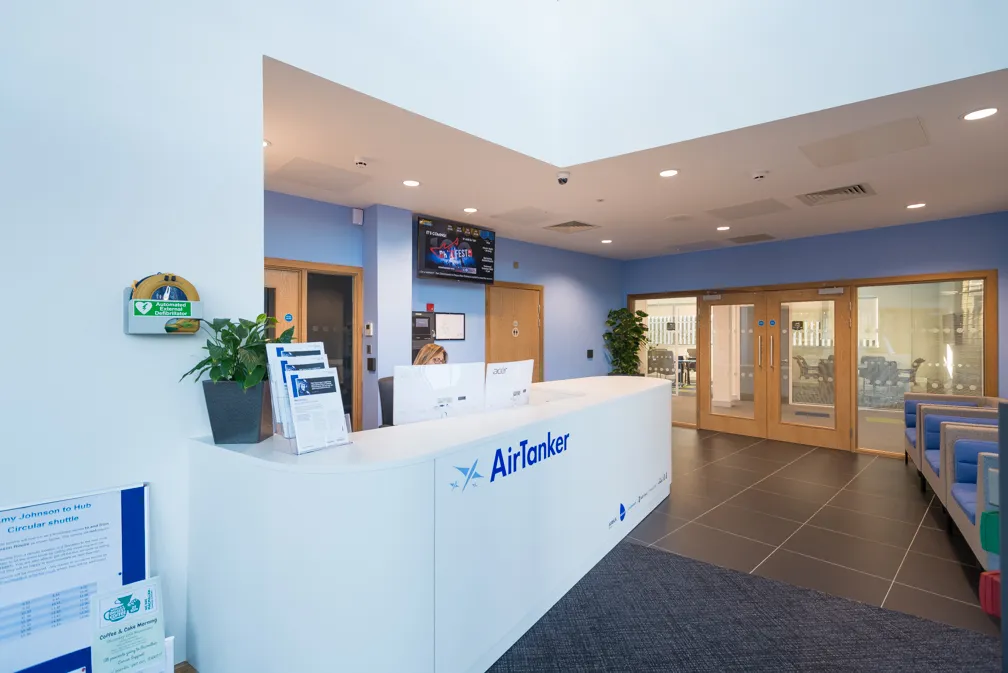 Modern office reception area with white AirTanker desk, computer monitors, potted plant, and seating area with blue chairs.