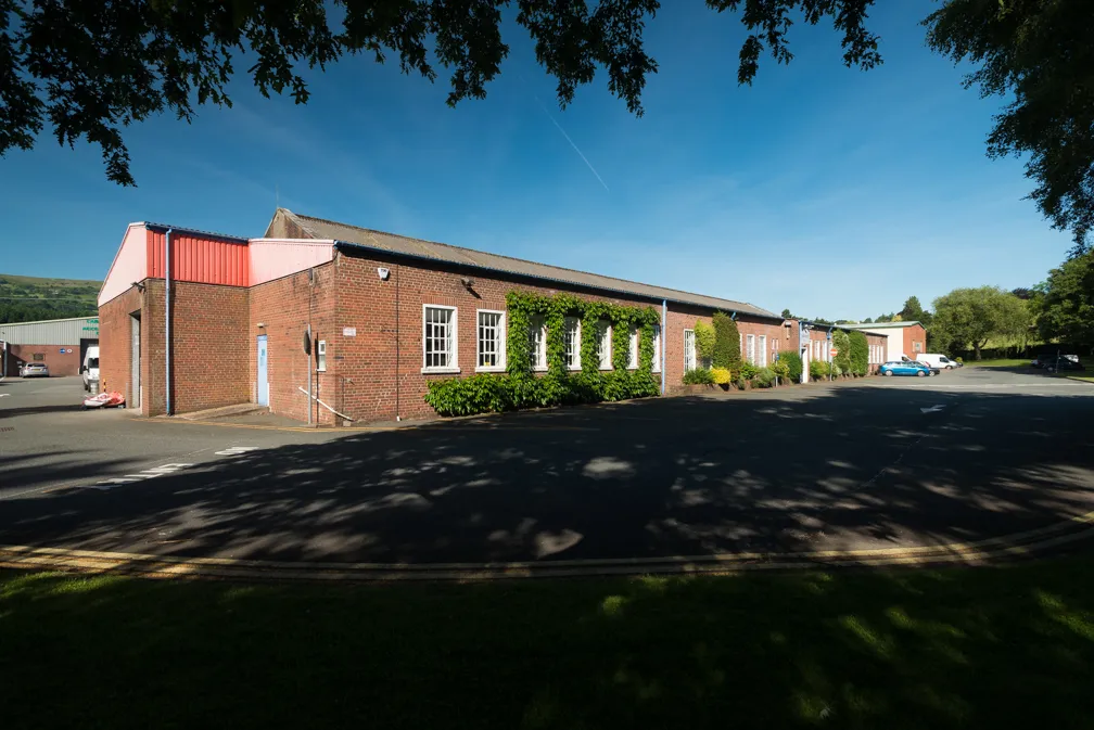 Long, single-story brick building with multiple windows and climbing plants, surrounded by a parking lot and trees under a clear blue sky.