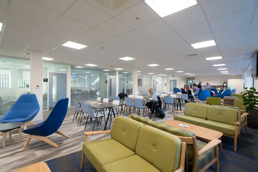 Modern office lounge with green sofas, blue armchairs, tables, and people working in the background.