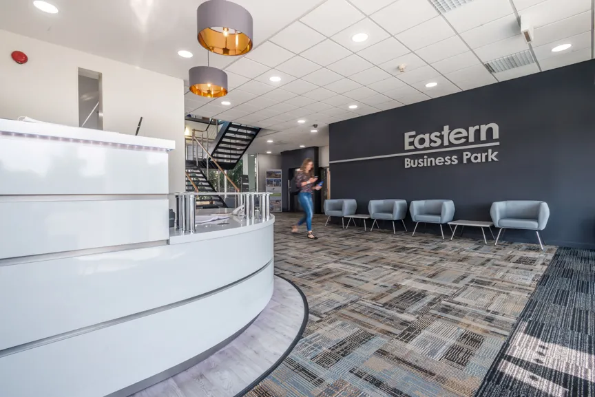 Modern business lobby with white curved reception desk, gray chairs along dark wall reading 'Eastern Business Park', and a staircase.