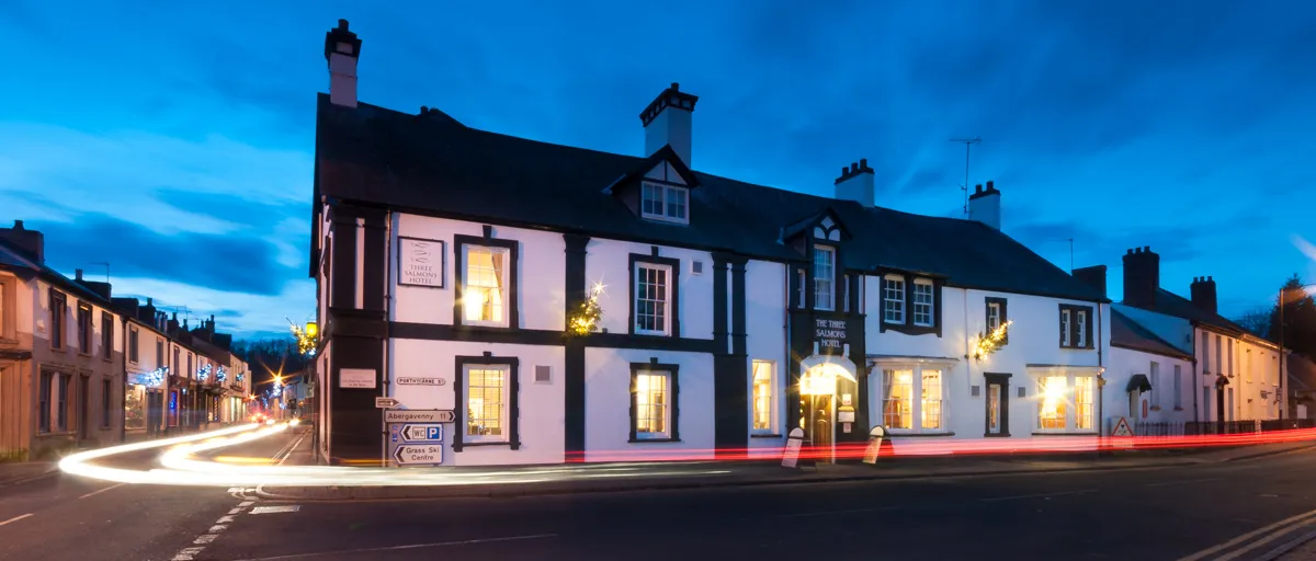 The Three Salmons Hotel building lit up at dusk with light trails from passing cars on the street corner.