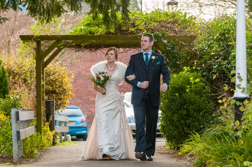 Bride in white dress with bouquet and groom in dark suit walking arm in arm under a garden pergola.