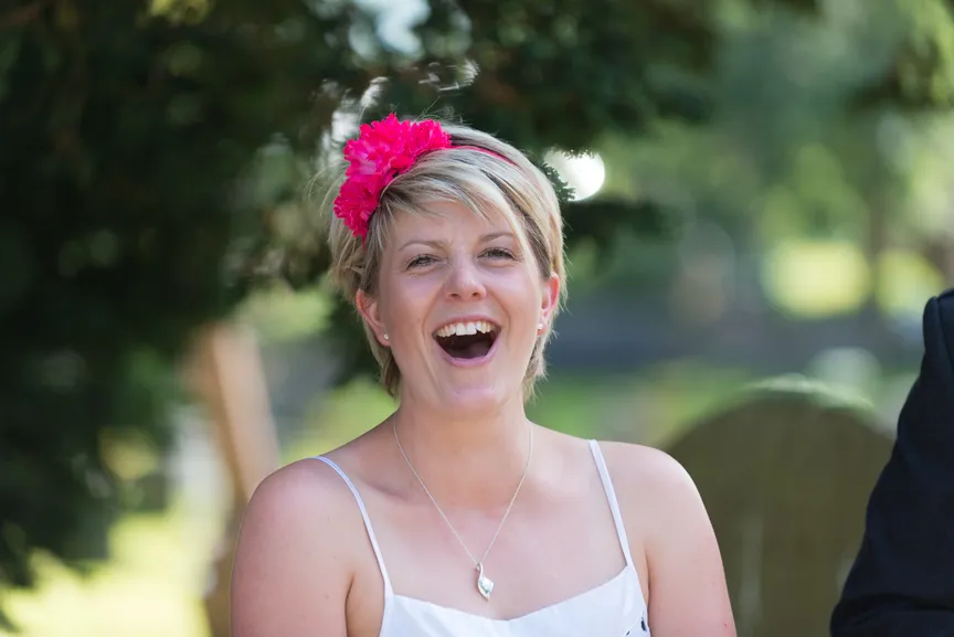 Smiling woman with short blonde hair wearing a white sleeveless top and a bright pink flower headband.