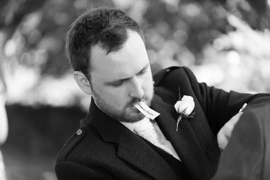 Man in a suit with a boutonnière holding a card labeled 'Grooms Father' in his mouth.