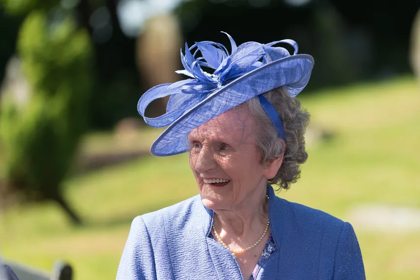 Smiling elderly woman wearing a blue decorative hat and matching jacket outdoors.