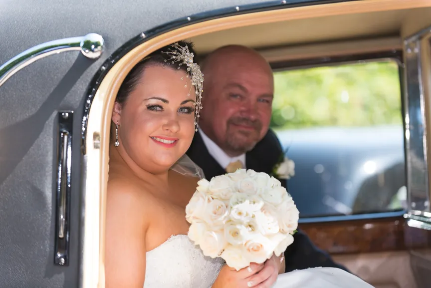 Smiling bride holding a bouquet of white roses sitting next to a groom inside a vintage car.