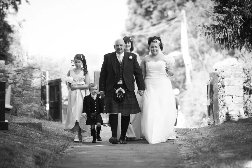 Black and white image of a bride in a wedding dress and groom in a kilt walking down a stone path accompanied by three attendants, including a young boy in traditional Scottish attire.