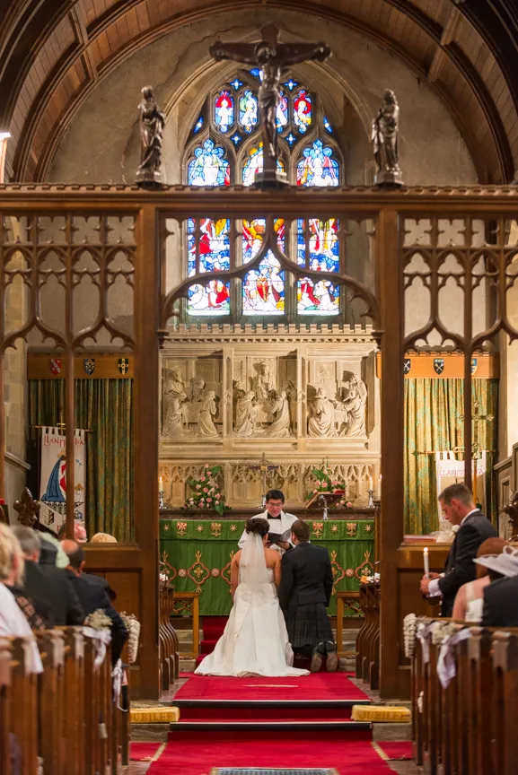 Bride and groom kneeling at the altar in a church during a wedding ceremony with stained glass windows and religious sculptures in the background.