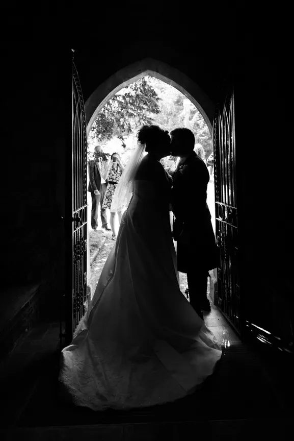 Black and white silhouette of a bride and groom kissing in a doorway with guests visible outside.