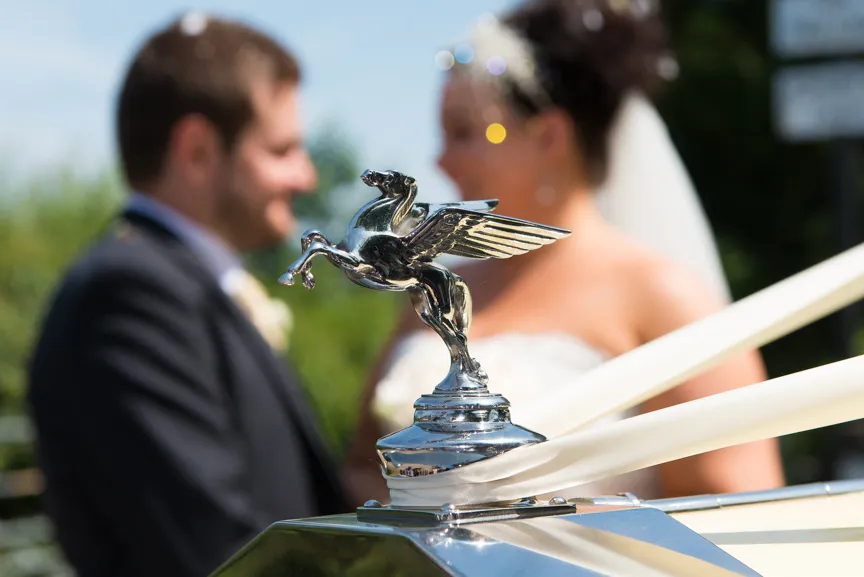 Silver winged horse hood ornament on a classic car with a blurred bride and groom in the background.