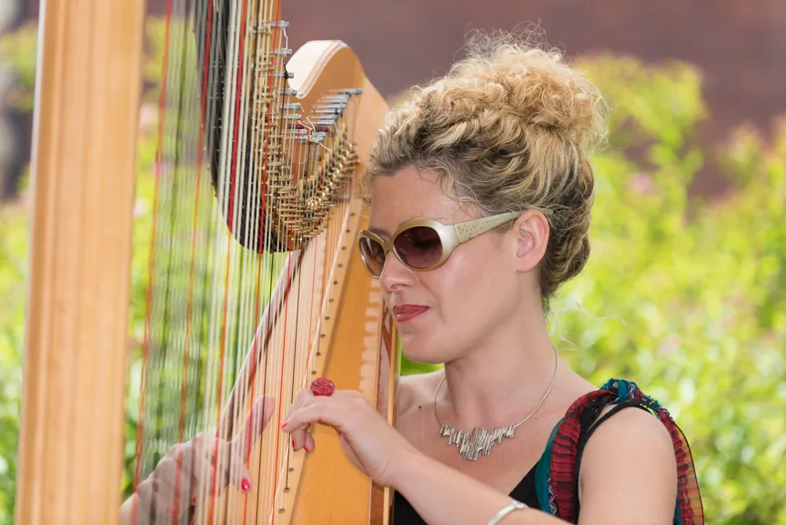 Woman with sunglasses playing a wooden harp outdoors with green foliage in the background.