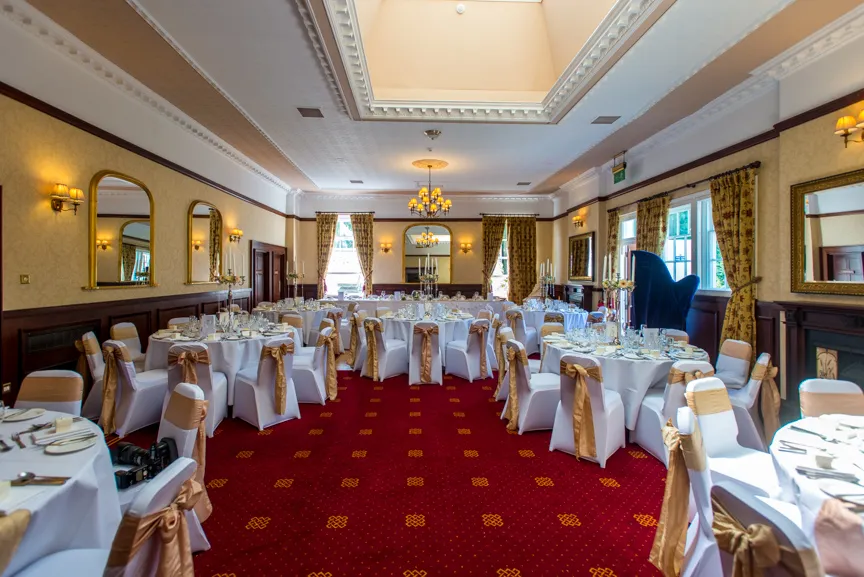 Elegant banquet hall with white chairs tied with gold sashes surrounding round tables set for an event under a skylight.
