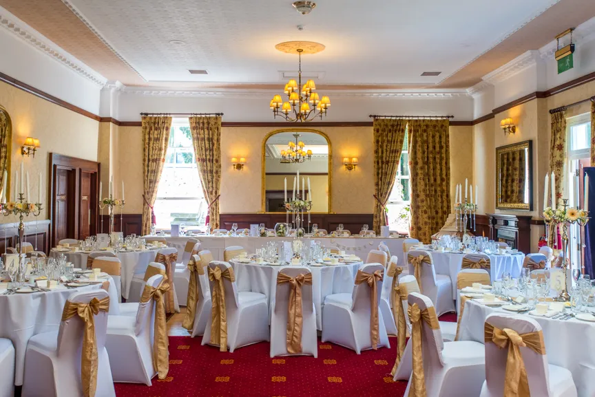 Elegant banquet hall with round tables covered in white cloths, chairs with gold sashes, chandeliers, and a large mirror on the wall.